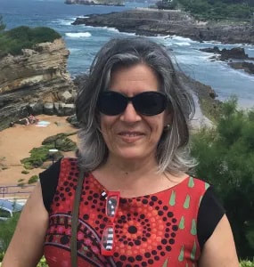 Foto retrato de mulher com cabelo pelos ombros, cinzento, com óculos de sol, sorrindo. Veste uma t-shirt vermelha com padrão preto e pormenores verdes. O fundo é uma praia.
